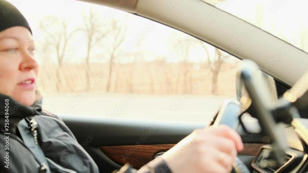 Warmly dressed female driver talking to a fellow traveler while driving car along highway with passing trees during the day in winter, traveling in nature. Road trip. Warmly dressed female driver talking to a fellow traveler while driving car along highway with passing trees during the day in winter, traveling in nature. Road trip.