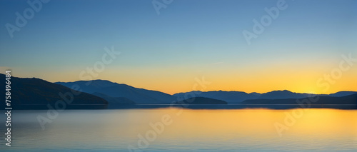Peaceful coast of the lake with the sun setting, its yellow glow mirrored perfectly on the lake’s surface, under a clear blue sky.