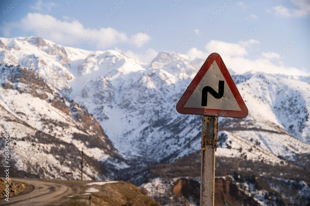 A winding mountain road disappears into the distance, framed by a cautionary road sign and the backdrop of snow-clad mountains. This image speaks to the careful attention needed when navigating such t A winding mountain road disappears into the distance, framed by a cautionary road sign and the backdrop of snow-clad mountains. This image speaks to the careful attention needed when navigating such t