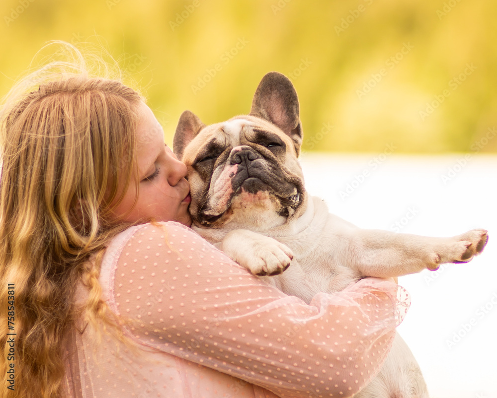 On a hot summer day outside, a beautiful, elegant girl is holding a French bulldog in her arms. A model girl and a dog. On a hot summer day outside, a beautiful, elegant girl is holding a French bulldog in her arms. A model girl and a dog.