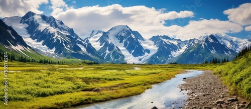 A river flows through a lush grassy field with mountains in the background, under a sky filled with fluffy clouds, creating a peaceful natural landscape