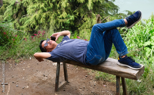 Man lying on old wooden bench over hilltop making a call on his smartphone photo