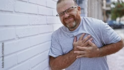 Handsome middle age caucasian man suffering heart attack outdoors on a sunny city street, emotion of pain evident as he clutches chest, leaning against wall with a look of sickened love.