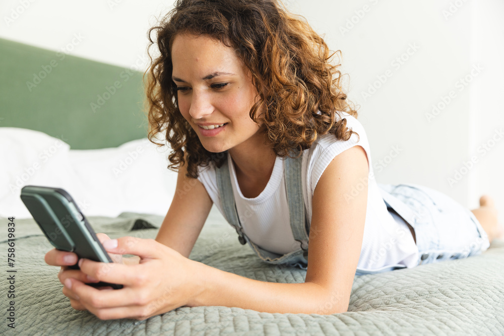 A young Caucasian woman is engrossed in her smartphone, relaxing on a bed at home A young Caucasian woman is engrossed in her smartphone, relaxing on a bed at home