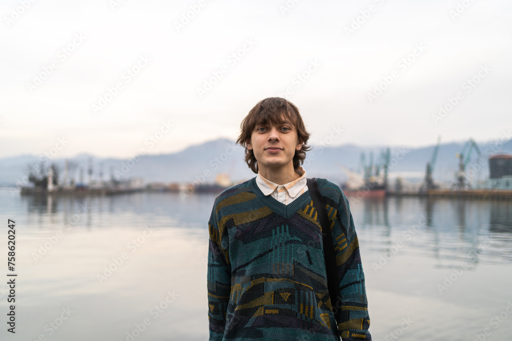 Young man in vintage inspired attire stands before tranquil harbor backdrop, fusing classic style with maritime setting.