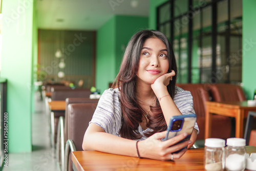 Free Download Thoughtful young Asian woman wearing dress sitting at a restaurant for a breakfast, holding a smartphone while propping her chin - GFXLoad