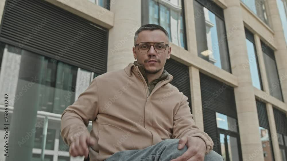 Zoom portrait shot of young man in wheelchair straightening his glasses and looking at camera with confidence in front of modern office building Zoom portrait shot of young man in wheelchair straightening his glasses and looking at camera with confidence in front of modern office building
