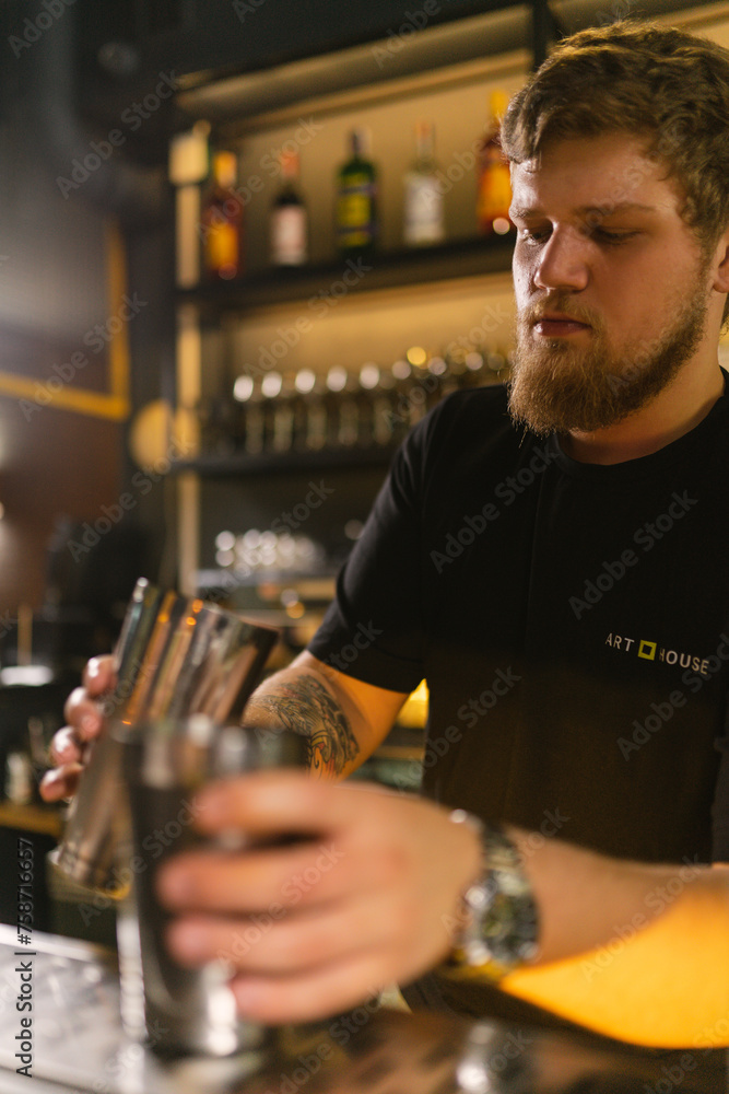 Young bearded bartender makes cocktail for customer. Skillful man holds shaker filled with ingredients standing by bar counter