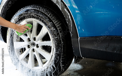A person is washing a car tire with a green brush. Tire is covered in water and soap. Blue car wash with white soap foam. Auto care service. Car cleaning service concept. Vehicle cleaning service.