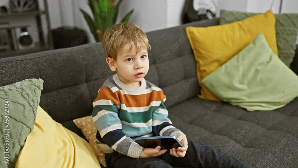 A focused toddler boy sits on a gray couch holding a smartphone, in a cozy living room setting. A focused toddler boy sits on a gray couch holding a smartphone, in a cozy living room setting.