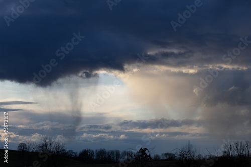 Gro  e Dunkle Wolke regnet etwas ab  im Hindergrund Sonne  blauer Himmel und wei  e und graue Wolken  im Vordergrund  schwarze Silhouetten von B  schen und B  umen 