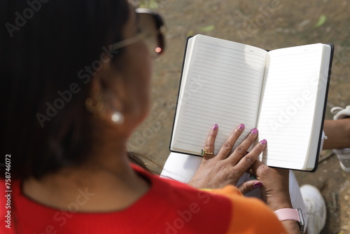 Portrait of mid aged woman holding a black color almanac in hand, reading personal almanac. sunlight, green park environment. book promotion, blank pages.