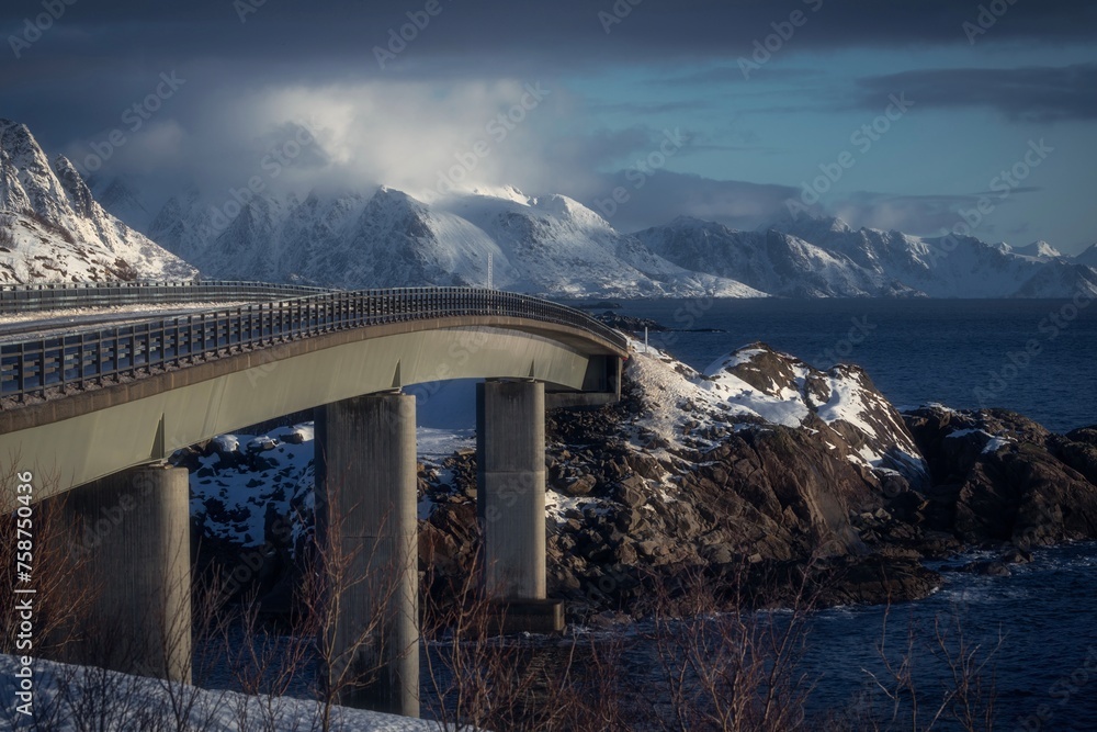 The image depicts a bridge spanning over water, with mountains in the background. The scenery includes a cloudy sky and a winter landscape with snow-capped mountains.

