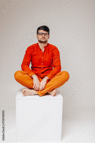 Man in glasses and bright orange clothes sits in the lotus position on a cube looking into the frame