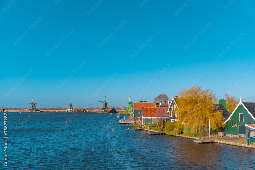 Naklejka premium Beautiful Dutch scenery panorama of Zaanse Schans village in Netherlands in spring time. Ancient windmills at the water's edge in the background with the blue sky in sunny weather