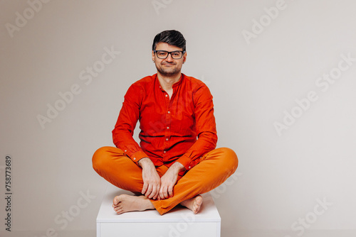 Man in orange clothes and glasses sits on a white cube and looks into the camera lens