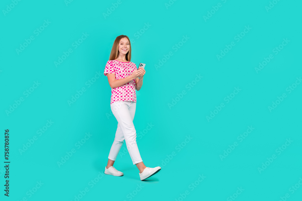 Full size photo of pretty teenager girl walk hold gadget eshop dressed stylish pink print outfit isolated on aquamarine color background Full size photo of pretty teenager girl walk hold gadget eshop dressed stylish pink print outfit isolated on aquamarine color background