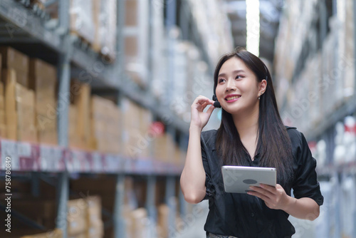 Asian attractive smile Female Inventory Manager check warehouse stock and talk Information to customer. They check product stock. In the Background Stock of Parcels with Products Ready for Shipment.