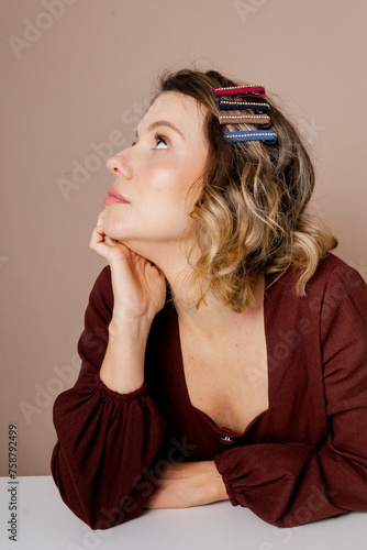 Bored young woman with dark blond hair and brown blouse on isolated beige background. Using hair accessories.