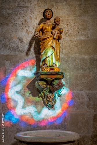 Virgin and child statue illuminated by the colorful light of a stained glass window in the church of Saint-Paul de Vence, France