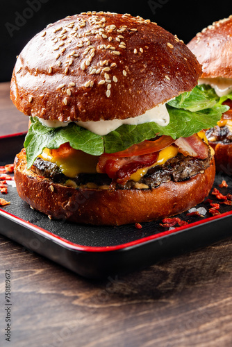 Close-up of a juicy burger with fresh lettuce, tomato, cheese, and a grilled patty on a sesame seed bun, served on a black plate. The background is dark, highlighting the vibrant colors of the burger