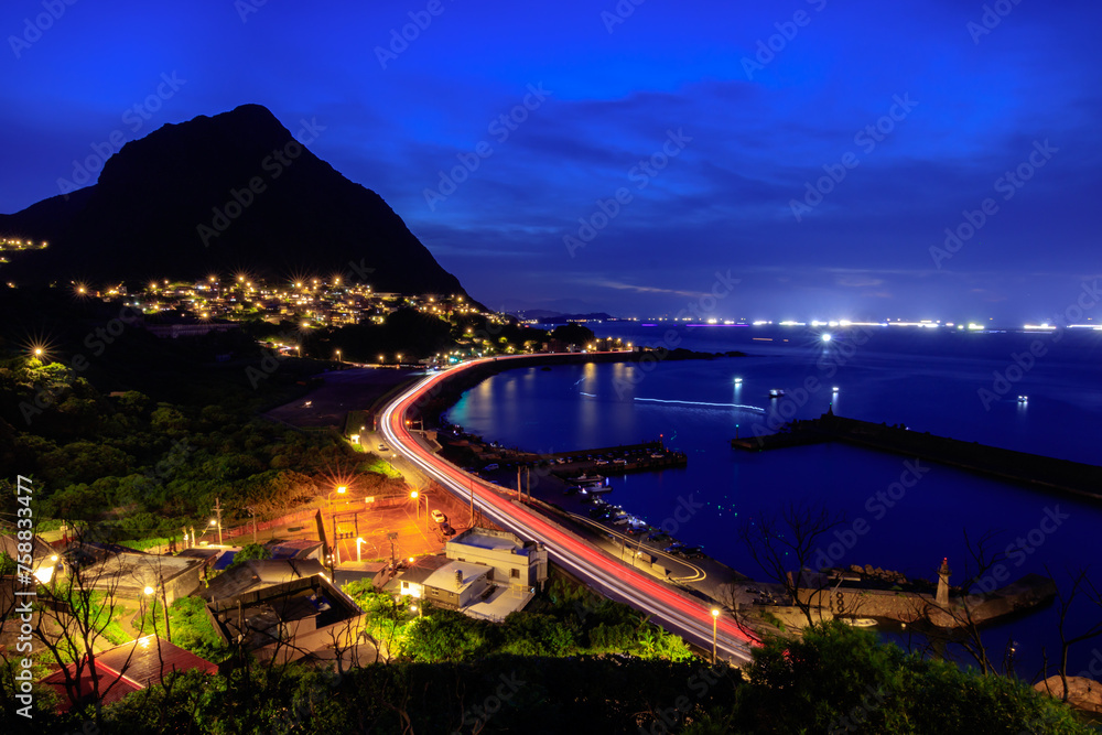 Light tracks of cars  along a c-shaped section of coastal highway in northern Taiwan accompanies with multiple scenery of sunset, steep mountains, village lights, a fishing port and quiet sea water.