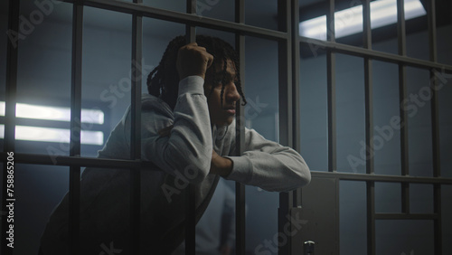 Cuadro en lienzo Upset African American teenager with face tattoos stands in prison cell in jail or youth detention center leaning on metal bars