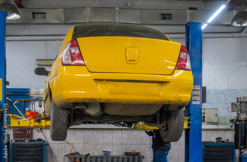 A yellow car being repaired hangs on a lift in a car repair shop. A yellow car being repaired hangs on a lift in a car repair shop.