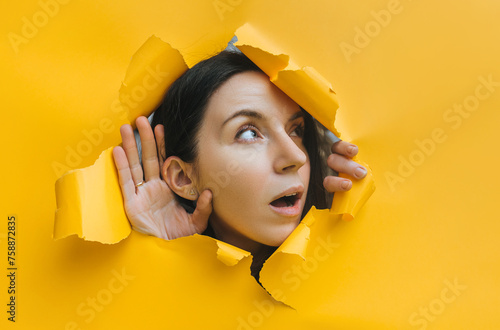A young girl puts her hand to her ear and listens in surprise, her mouth open through a hole in the paper. Bright background, copy space. The concept of eavesdropping,espionage,gossip and yellow press