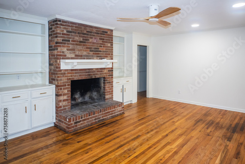 A living room or den with hardwood floors, white walls and a red brick fireplace with mantle in a newly renovated house