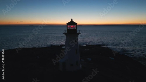 Cinematic drone shot Coastal lighthouse, Peggy's Cove Lighthouse against sunset sky at dusk, Atlantic Coast Lighthouse Halifax Nova Scotia Canada. The light comes from a first order Fresnel lens.  photo
