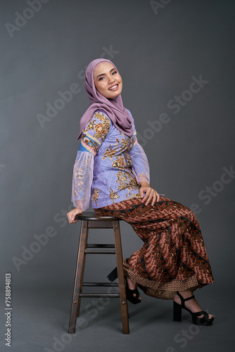 Beautiful female model wearing modern batik kebaya,  sitting on a chair isolated over grey background. Eidul fitri festival fashion and beauty concept.