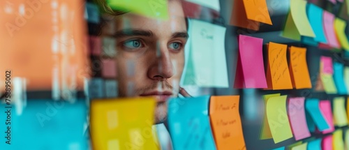Male programmer looking at adhesive notes posted on the window of his start-up office in a thoughtful manner