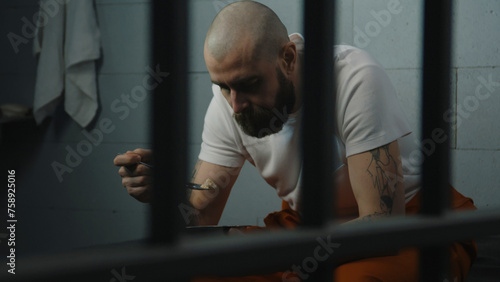 Inmate in tattoos in orange uniform sits on bed in jail cell, eats prison food from iron bowl. Male prisoner serves imprisonment term for crime in prison. Detention center or correctional facility. photo