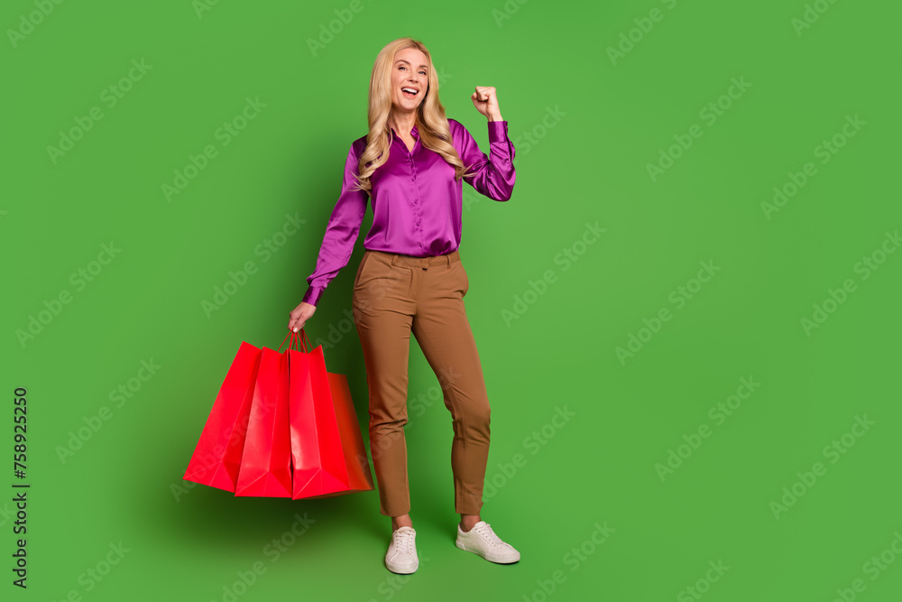 Full size photo of delighted gorgeous lady hold store bags raise fist empty space isolated on green color background Full size photo of delighted gorgeous lady hold store bags raise fist empty space isolated on green color background