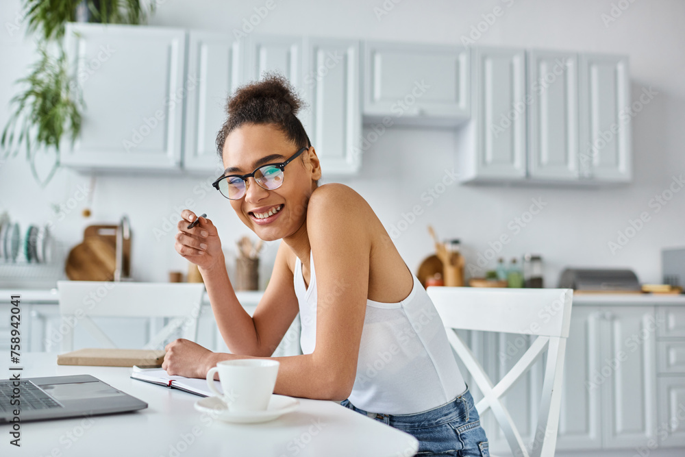 cheerful african american woman in glasses taking notes near laptop and cup of coffee, remote work cheerful african american woman in glasses taking notes near laptop and cup of coffee, remote work