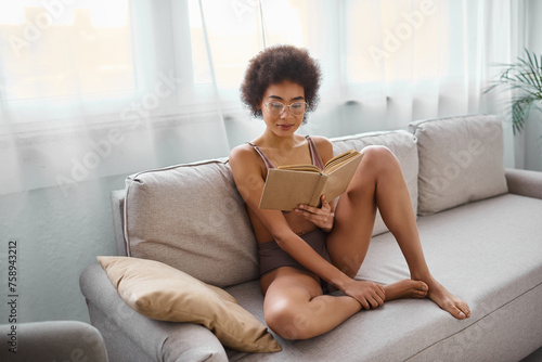 relaxed and curly-haired african american woman reading novel in lingerie on a comfy sofa