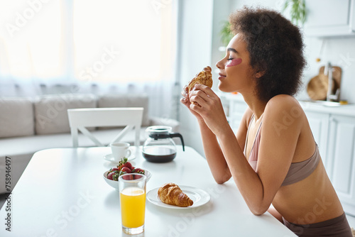 smiling african american woman holding freshly baked croissant during breakfast in kitchen