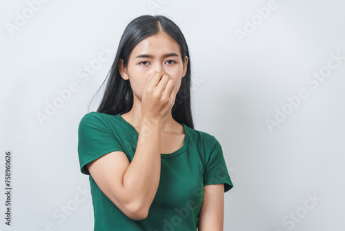 Young Asian frustrated woman with green t-shirt pinches her nose with disgust on her face, reacting to a bad smell in isolation over background.