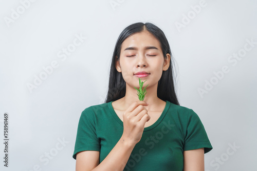 Young pretty Asian woman in a green t-shirt inhales the scent of the herbal rosemary isolated over white background.