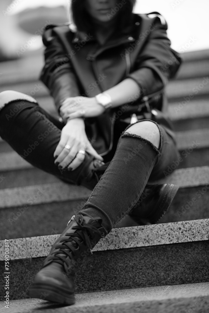 Fototapeta premium A beautiful girl with a stylish bob haircut is walking around the city, sitting on the stairs. The woman is wearing a black leather, sweater, torn jeans and boots