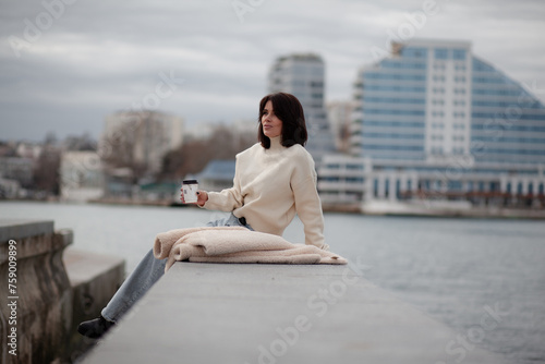 A beautiful girl with a stylish bob haircut is walking around the city, drinking coffee or tea, sitting against the sea. The woman is wearing a black leather, sweater, torn jeans and boots © Вероника Зеленина