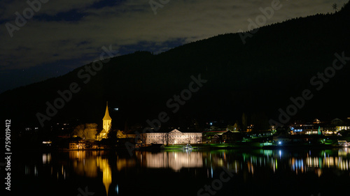 Noc. Oświetlony kościół z wieżą. Odbicia w ciemnej toni jeziora. Ciemny zarys góry. Alpy.