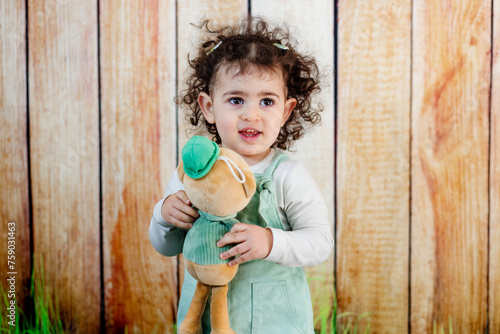 Obraz na płótnie A curly hair girl s hands wrapped around a plush friend, showcasing a loving bond in front of rustic wooden setting w salonie