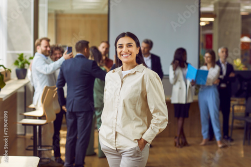 Portrait of a happy smiling confident young business woman looking at the camera with a team of company employees talking and working in the background in modern office after meeting.