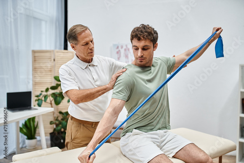 handsome patient in casual attire stretching with resistance band in front of his mature doctor © LIGHTFIELD STUDIOS