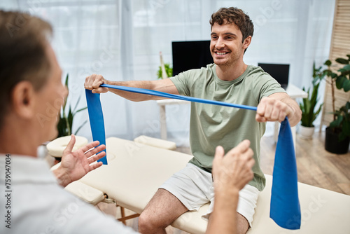 focus on jolly handsome patient using resistance band and smiling at his blurred mature doctor