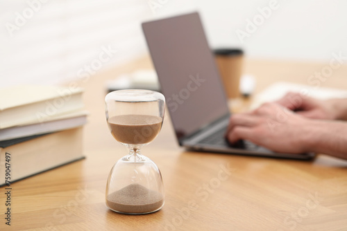 Hourglass with flowing sand on desk. Man using laptop indoors, selective focus