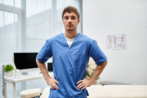 good looking rehabilitologist in blue medical robe posing in hospital ward and looking at camera
