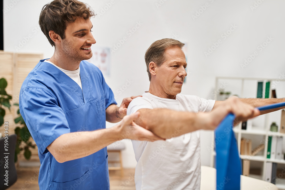 cheerful handsome doctor in blue robe helping his mature patient to use resistance band in hospital cheerful handsome doctor in blue robe helping his mature patient to use resistance band in hospital